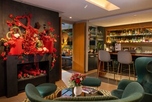 Una zona de bar con mostrador de madera y taburetes, una pared decorada con arreglos florales rojos y un área de asientos con sillas tapizadas de verde.