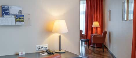 A hotel room with a desk, a telephone, a lamp, a chair, and a dining area with chairs and a table.