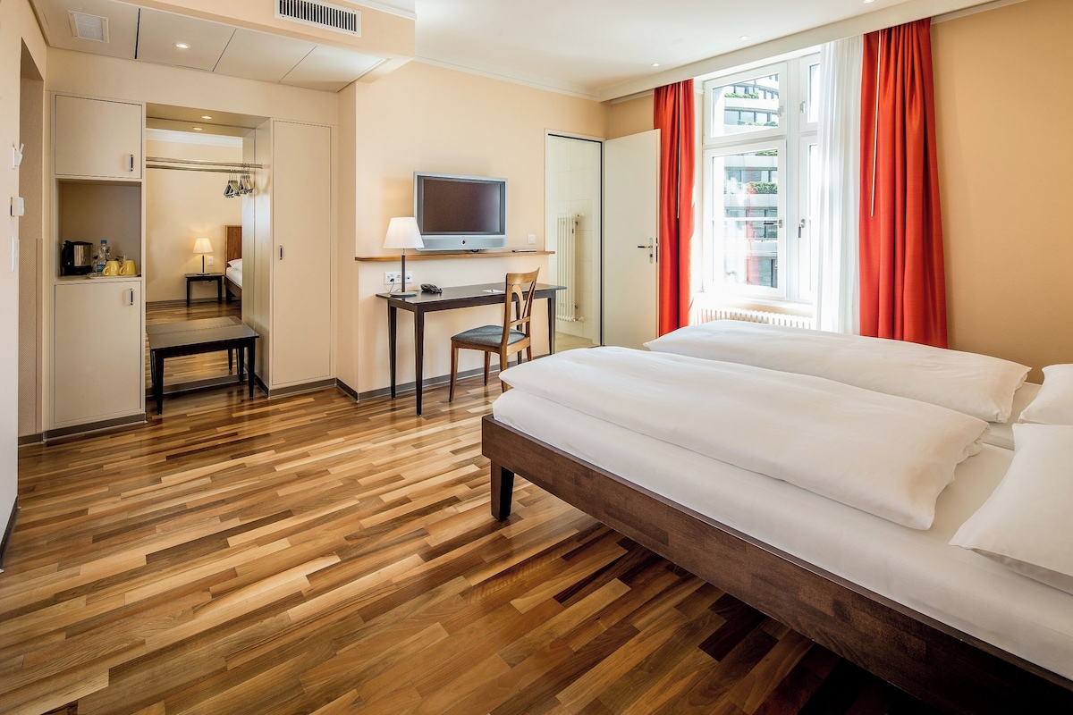 A hotel room with a wooden floor, a bed, a desk with a chair, a television, and a window with curtains.