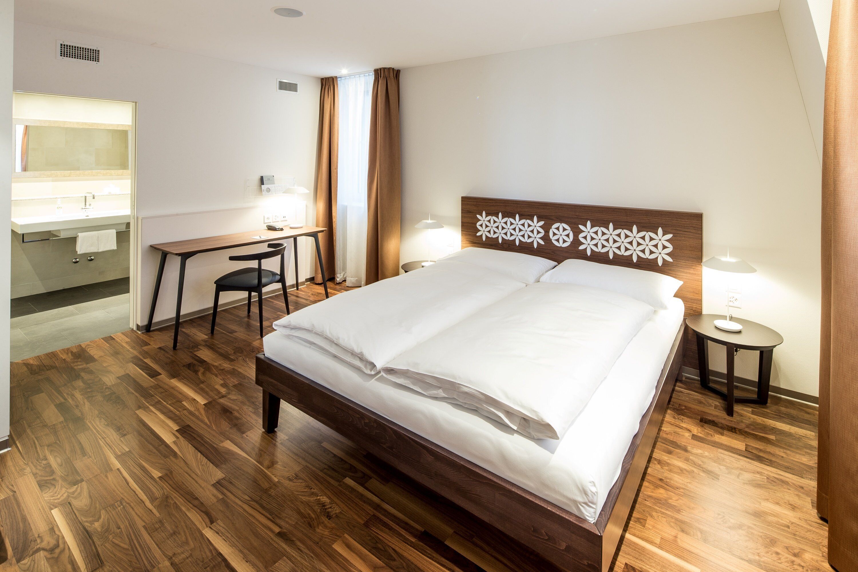 Ein Hotelzimmer mit Holzboden, einem Bett mit weißer Bettwäsche, einem Schreibtisch mit Stuhl, einem kleinen Tisch mit Lampe und einem Badezimmer mit Spiegel und Handtuchhalter.