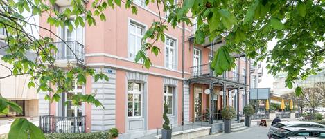 A pink building with a balcony, a parking area, and a few potted plants.