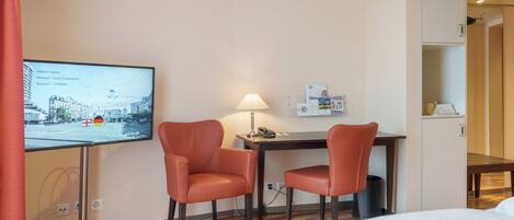 Una habitación de hotel con una cama, dos sillones naranjas, un escritorio con una lámpara, un televisor mostrando un paisaje urbano y un mueble pequeño.