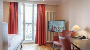 Une chambre d’hôtel avec un lit, un bureau avec une lampe, une chaise, une télévision et une fenêtre avec des rideaux.