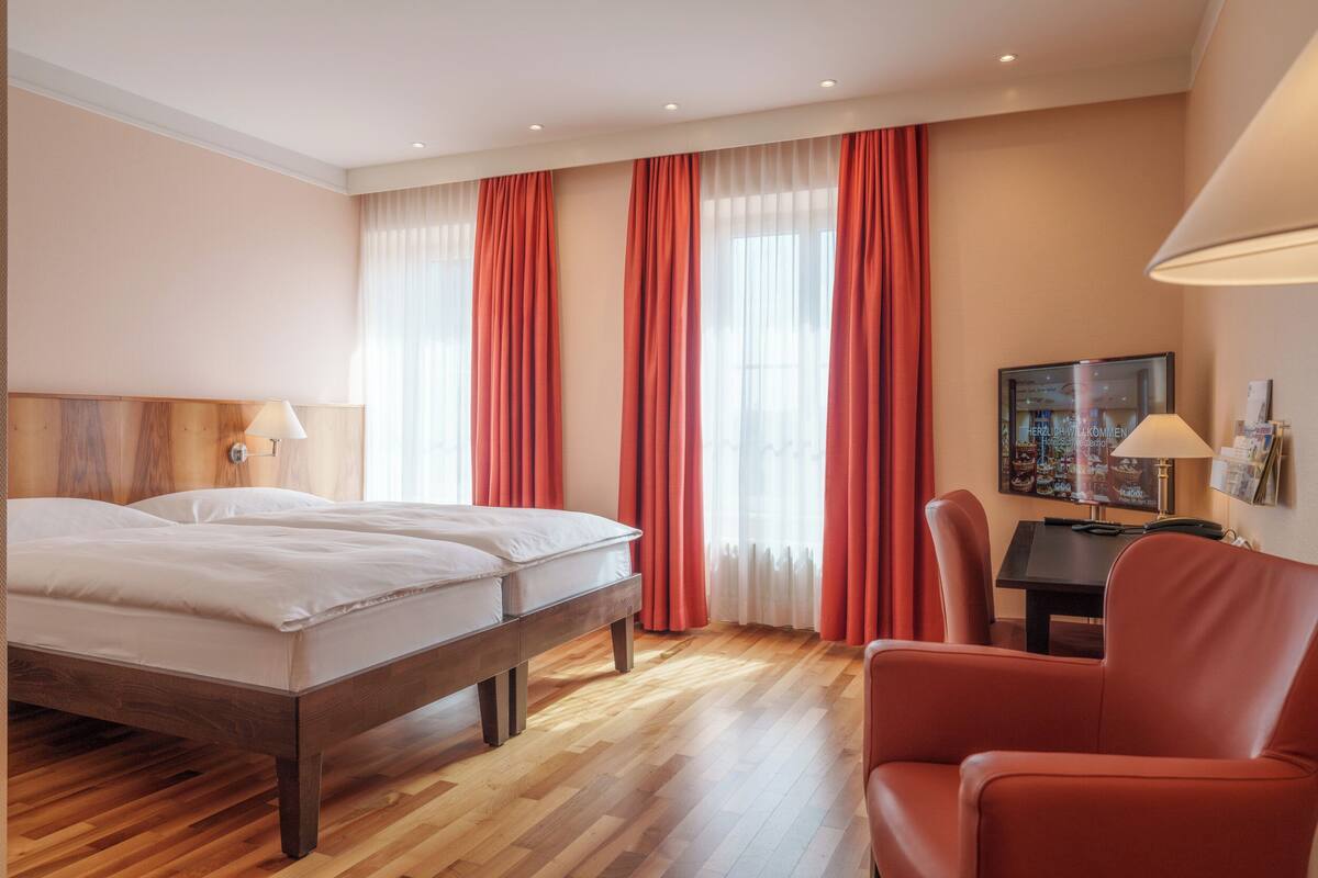 A hotel room with a wooden bed, red curtains, a desk with a lamp, and a chair.
