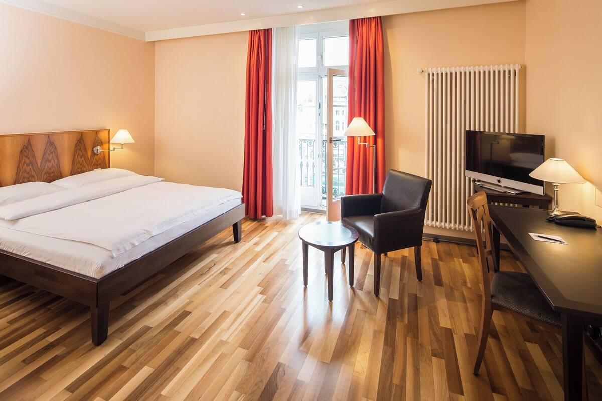A hotel room with a large bed, a desk with a TV, a chair, and a small table.