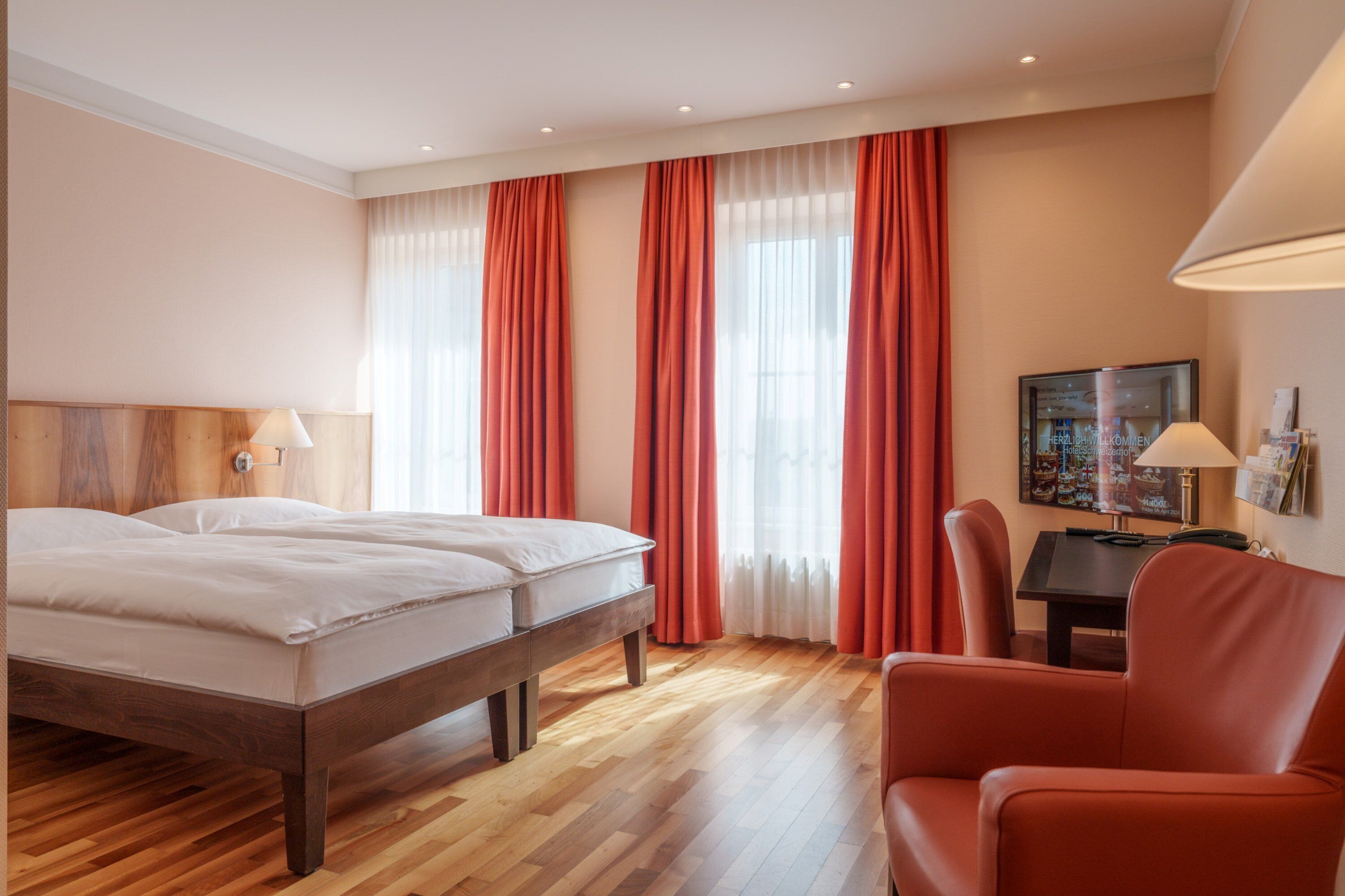 Una habitación de hotel con piso de madera, una cama con ropa blanca, una silla roja, un escritorio con una lámpara y un televisor en la pared.