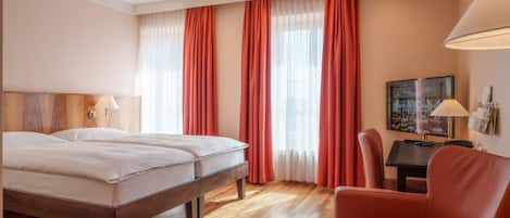 Una habitación de hotel con piso de madera, una cama con ropa blanca, una silla roja, un escritorio con una lámpara y un televisor en la pared.