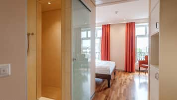 Una habitación de hotel moderna con cabina de ducha de vidrio, cama con cabecero de madera y vistas a la ciudad a través de amplios ventanales.