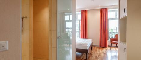 Una habitación de hotel moderna con cabina de ducha de vidrio, cama con cabecero de madera y vistas a la ciudad a través de amplios ventanales.