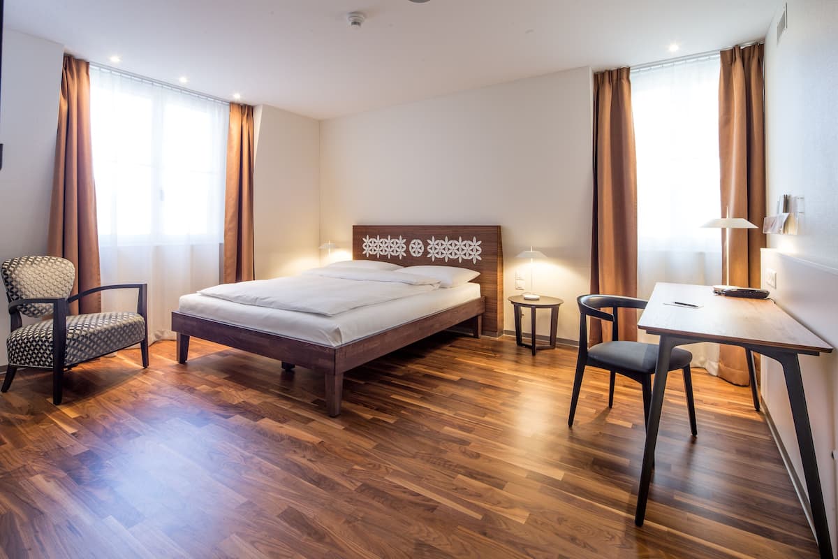 A hotel room with a wooden floor, a bed with a headboard, a desk with a chair, and two lamps.