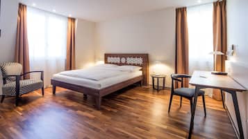 A hotel room with a wooden floor, a bed with a headboard, a desk with a chair, and two lamps.