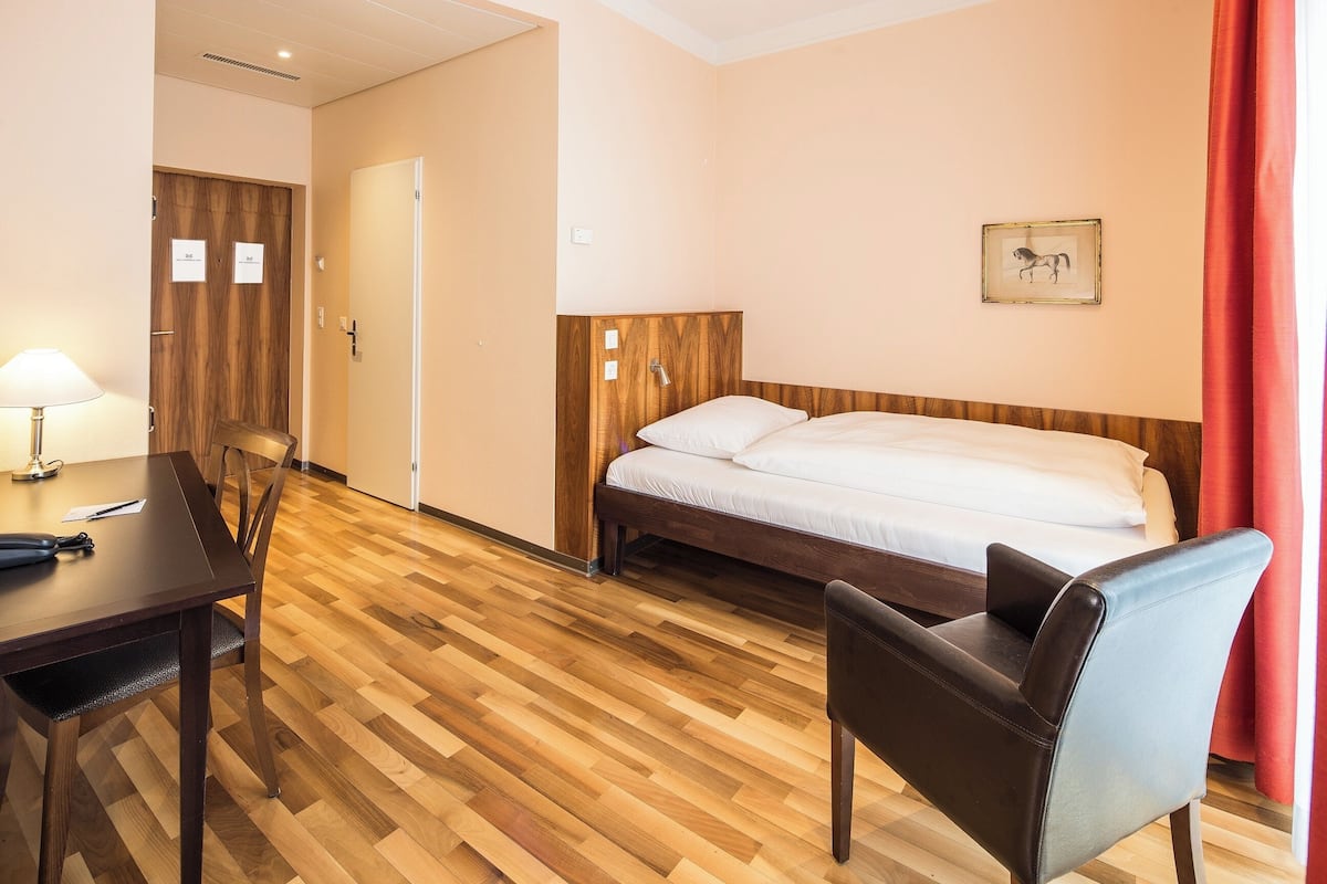 A hotel room with a wooden floor, a bed with a wooden headboard, a desk with a lamp, and a chair.