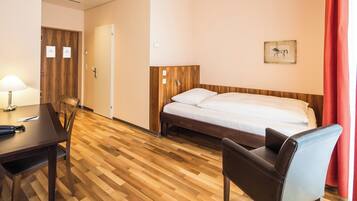 A hotel room with a wooden floor, a bed with a wooden headboard, a desk with a lamp, and a chair.