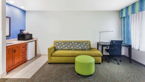 A compact room with a sofa, a small green ottoman, a desk with a chair, a microwave, and a refrigerator.