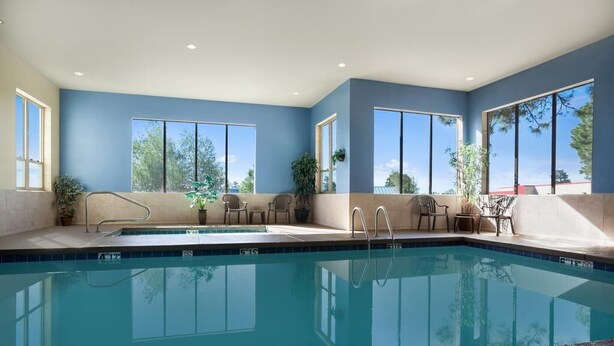 A modern indoor swimming pool with clear blue water, large windows, and a seating area with chairs and potted plants.