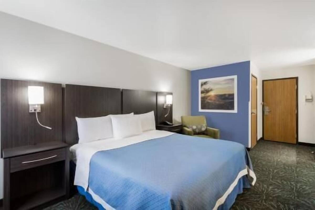 Habitación de hotel con cama, mesitas de noche, una silla, un cuadro en la pared y una puerta.