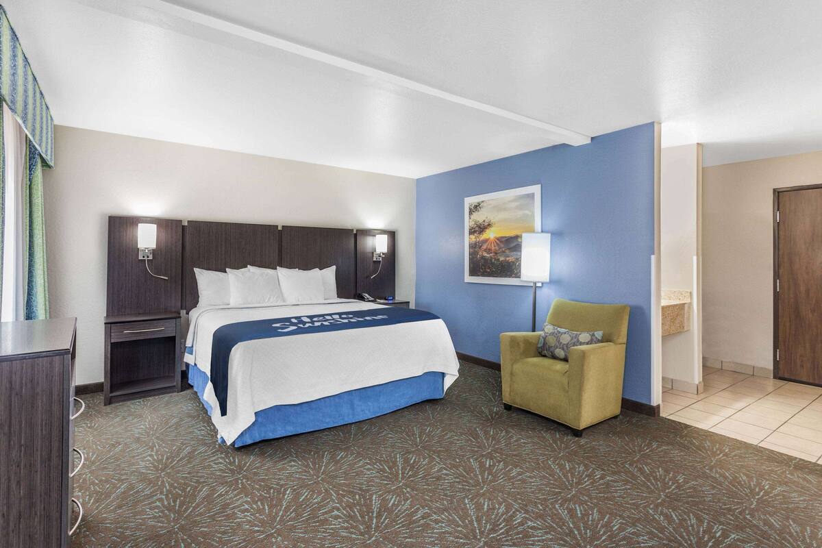 Una habitación de hotel con cama, mesita de noche, silla, lámpara y un cuadro en la pared.