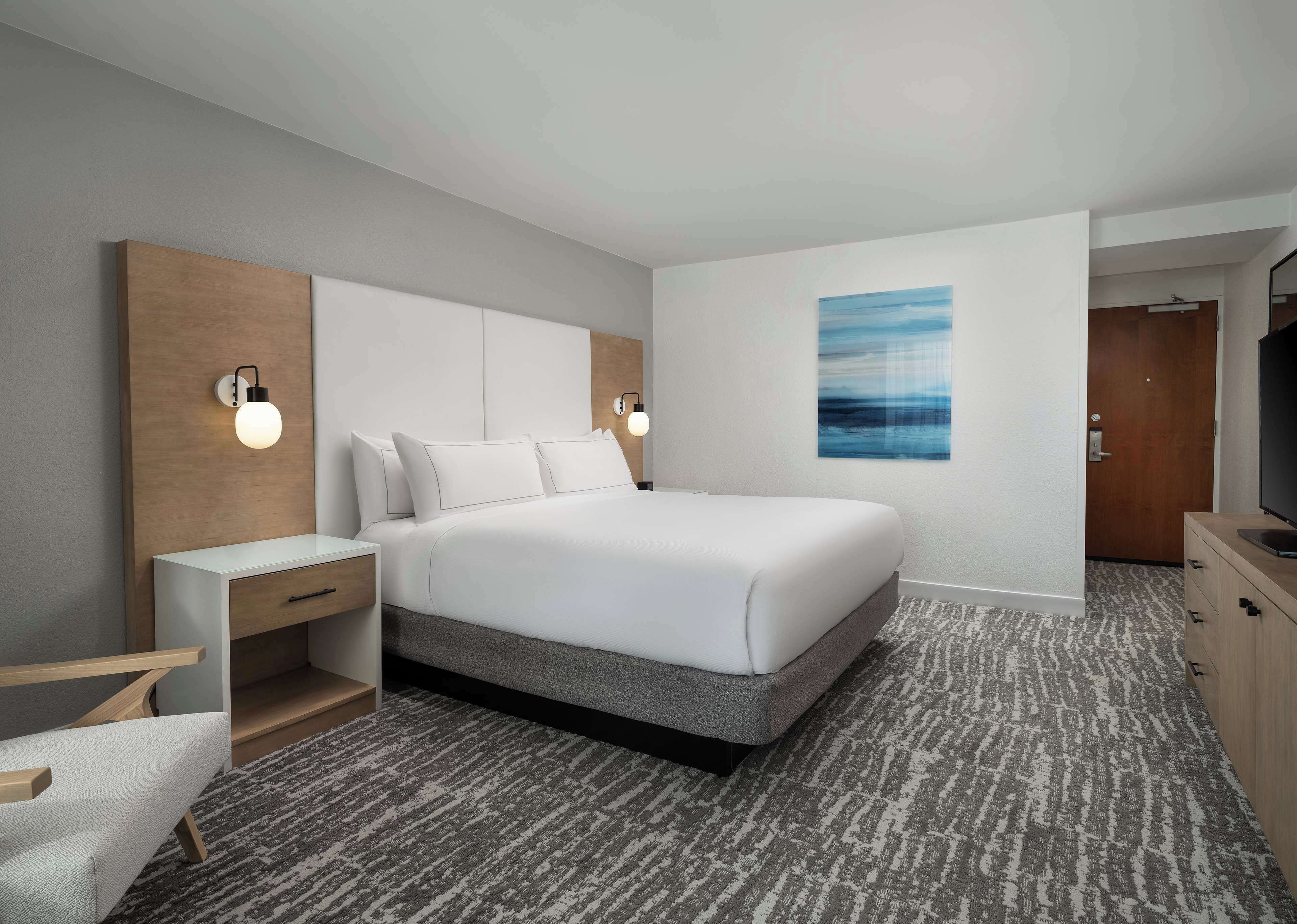 A modern hotel room with a large bed, bedside tables, a TV, and a painting on the wall.