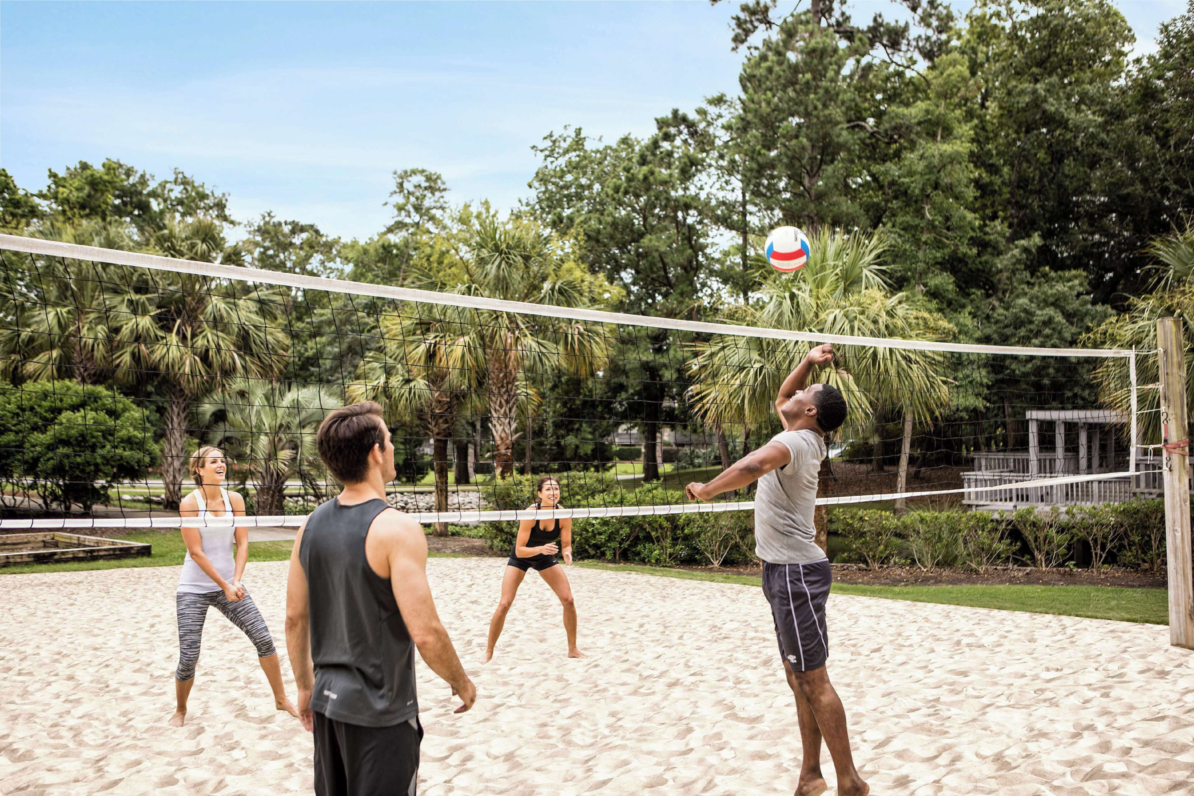 Ein Beachvolleyballspiel auf einem Sandplatz, umgeben von viel Grün.