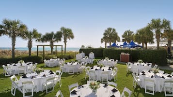 An outdoor wedding venue with round tables and chairs, a view of the ocean, and palm trees.