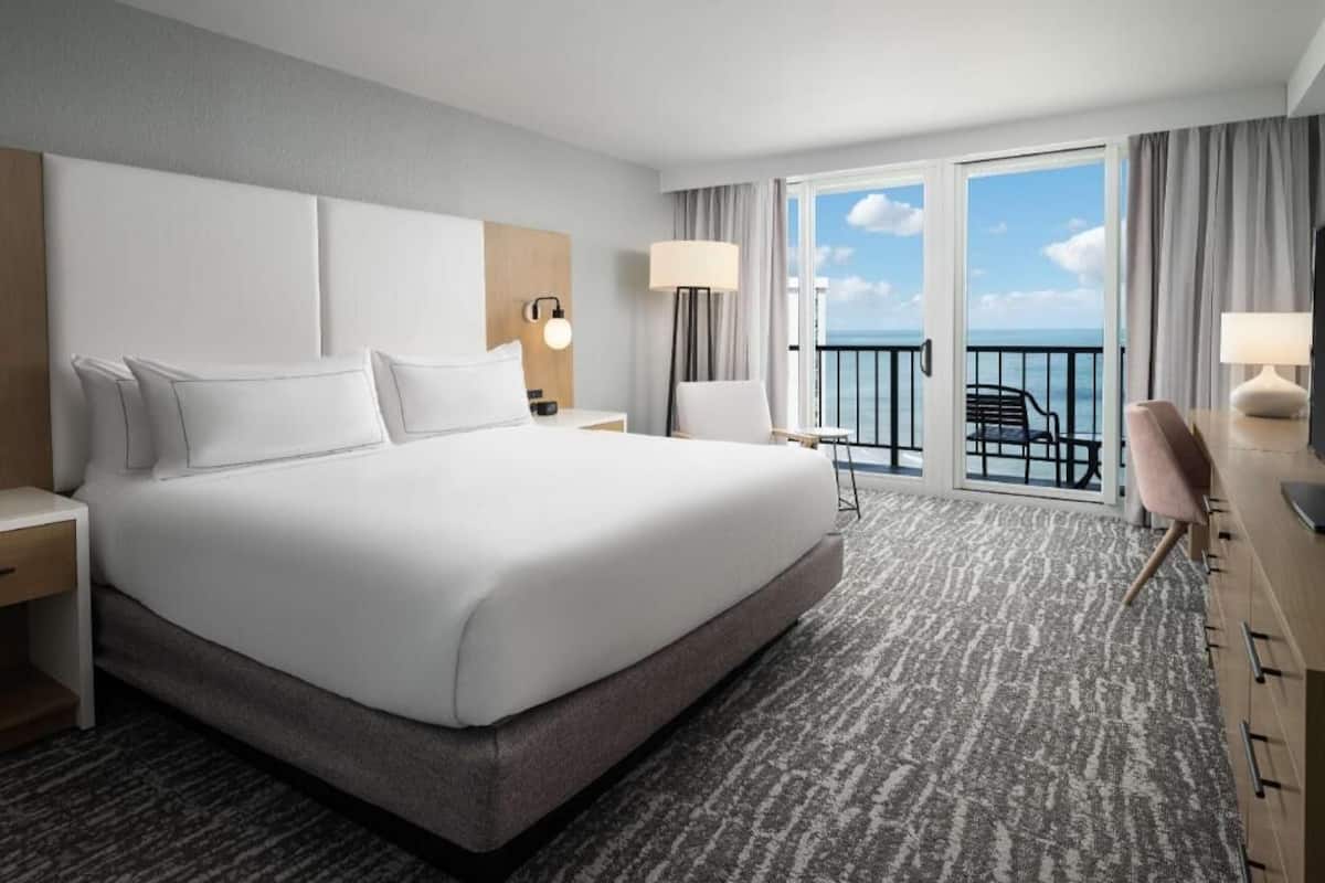 A hotel room with a large bed, a desk, a chair, a balcony with a view of the ocean, and a window with curtains.