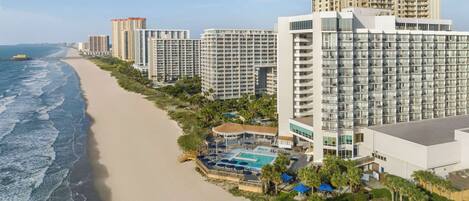 Un hotel frente a la playa con varios edificios altos, una playa de arena y una zona de piscina.