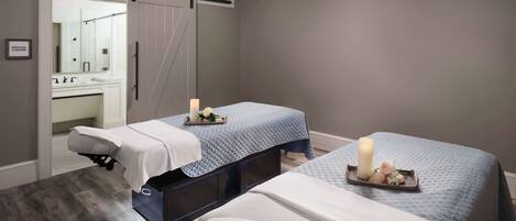 A spa room with two beds, a massage table, and a bathroom with a sink and mirror.
