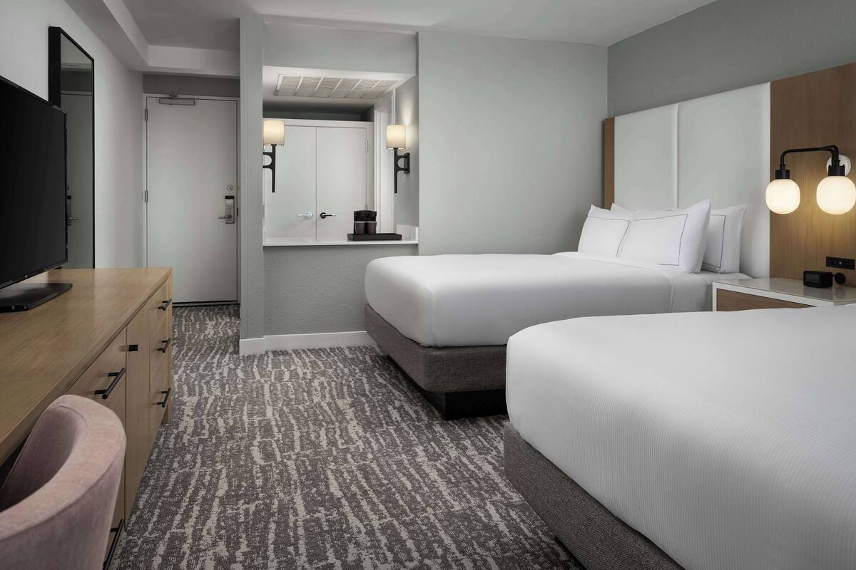 A hotel room with two beds, a desk with a television, a bathroom with a mirror, and a patterned carpet.