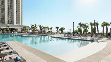 A swimming pool with lounge chairs and umbrellas, surrounded by palm trees and a tall building in the background.