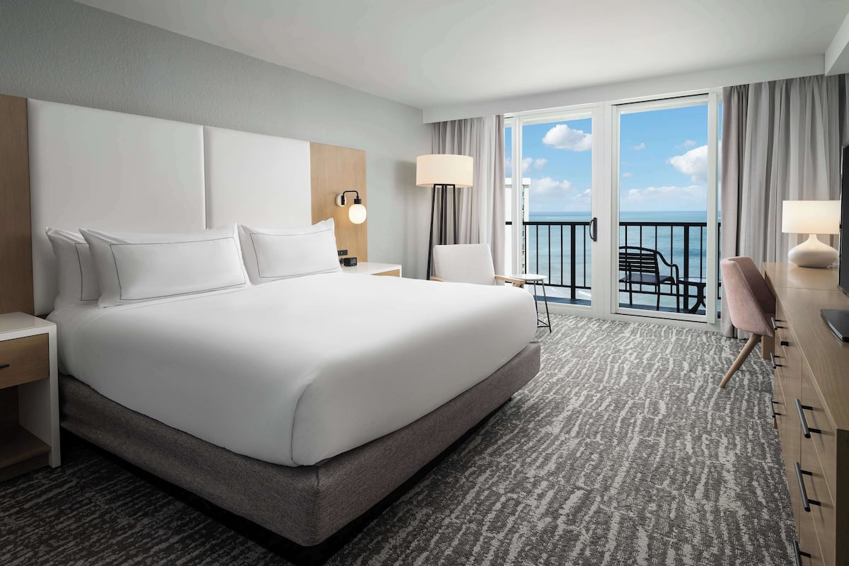 A hotel room with a large bed, a desk with a chair, a balcony with a view of the sea, and a TV.