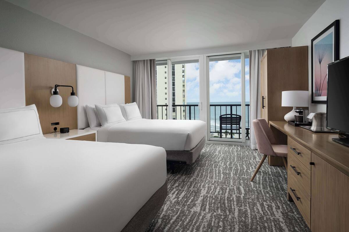 A hotel room with two beds, a desk, a chair, a TV, and a balcony with a view of the ocean.