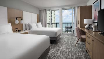 A hotel room with two beds, a desk, a chair, a TV, and a balcony with a view of the ocean.