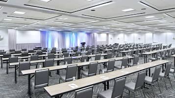 A conference room with rows of tables and chairs, a podium, and a large screen at the front.