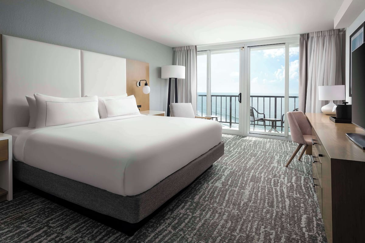 A hotel room with a large bed, a desk with a chair, a small table, and a balcony with a view of the sea.