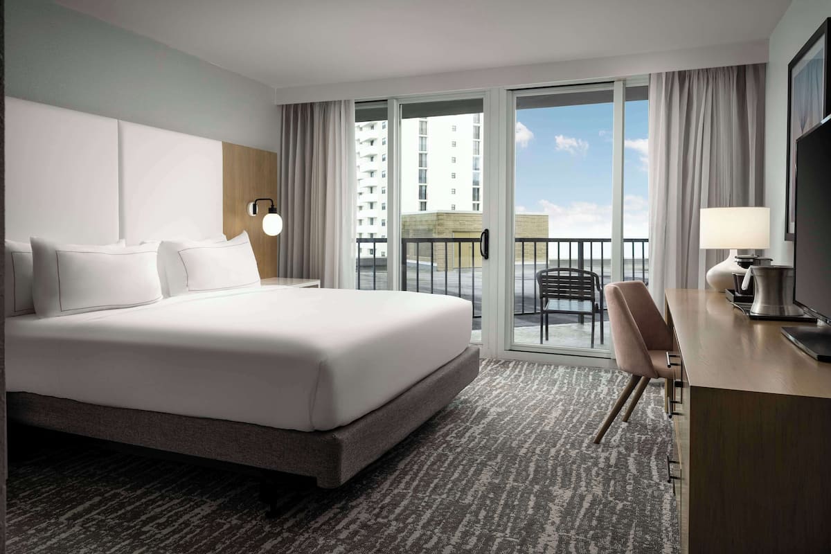 A hotel room with a large bed, a desk with a lamp, a chair, and a view of a building through the window.