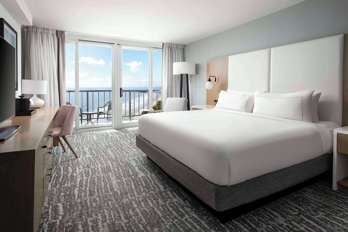 A hotel room with a large bed, a desk, a chair, a nightstand, and a view of the beach.