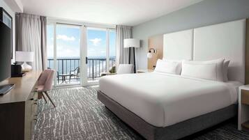 A hotel room with a large bed, a desk, a chair, a nightstand, and a view of the beach.