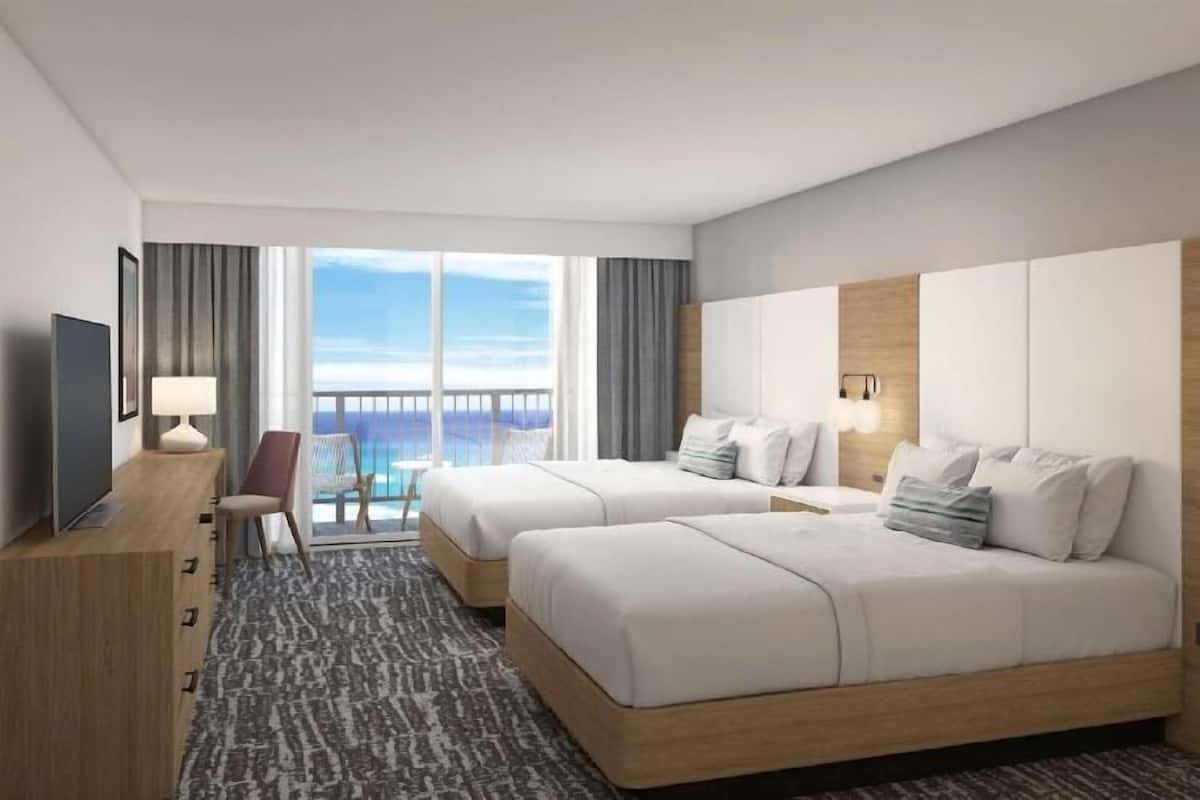A hotel room with two beds, a TV, a desk, and a balcony with a view.