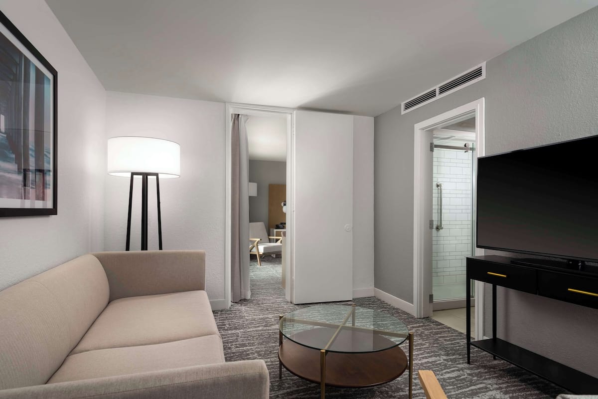 A hotel room with a sofa, a glass coffee table, a TV on a stand, a floor lamp, and a door leading to a bathroom.