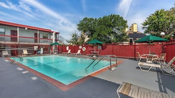 A swimming pool with lounge chairs and umbrellas, surrounded by a red fence, with a building in the background.