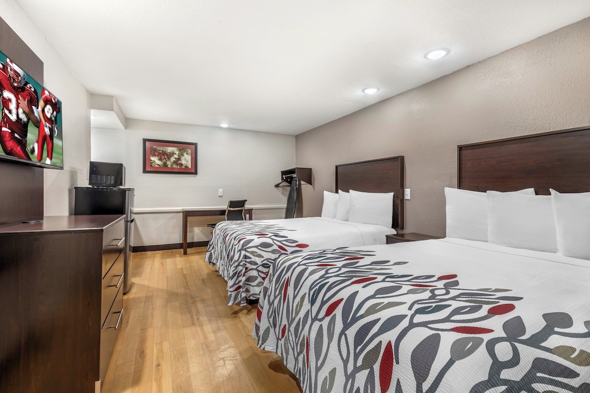 A hotel room with a long bed, a desk, a chair, a television, and a framed picture on the wall.