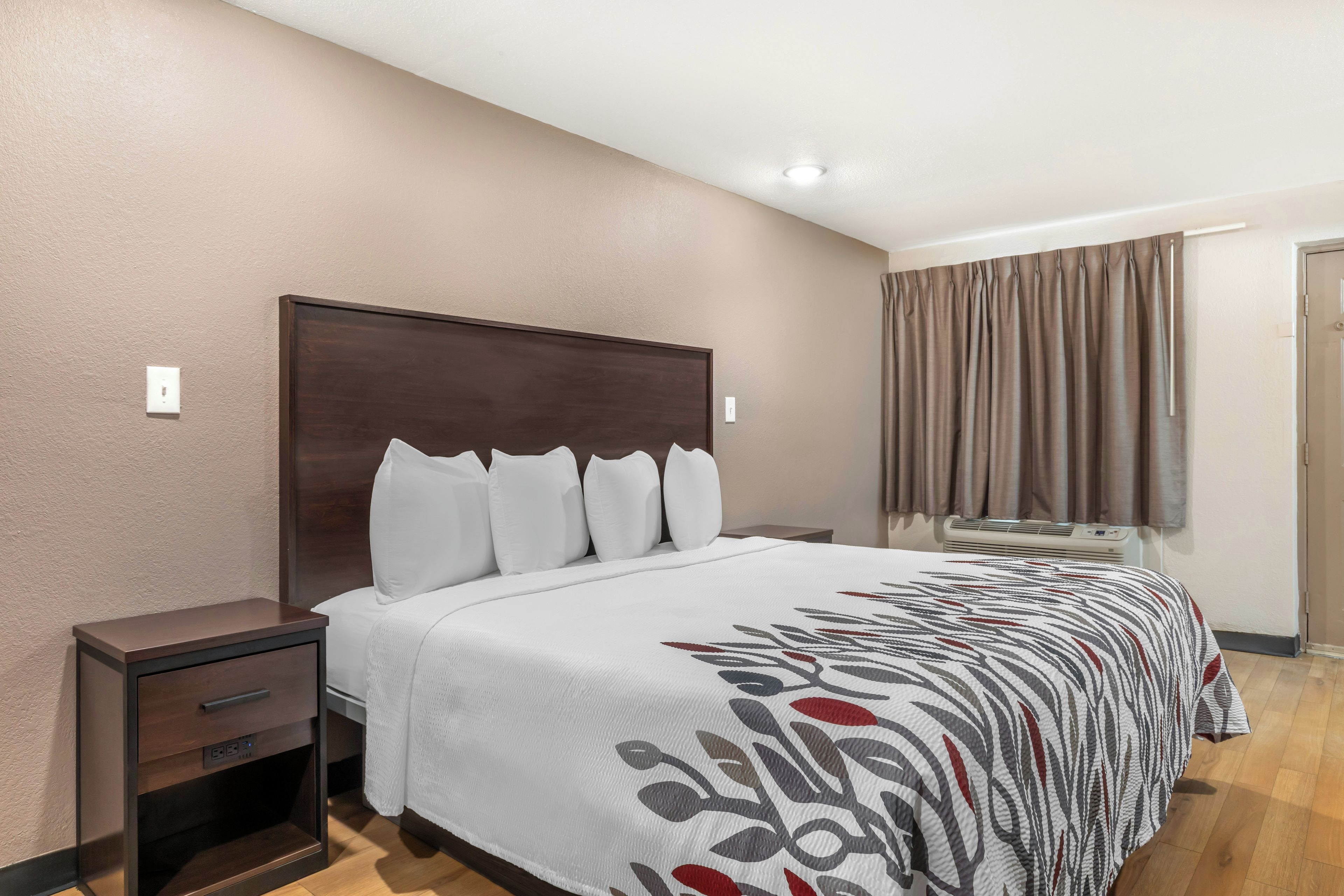 Habitación de hotel con una cama grande, una mesita de noche, un cajón, una ventana con cortinas y un equipo de aire acondicionado.