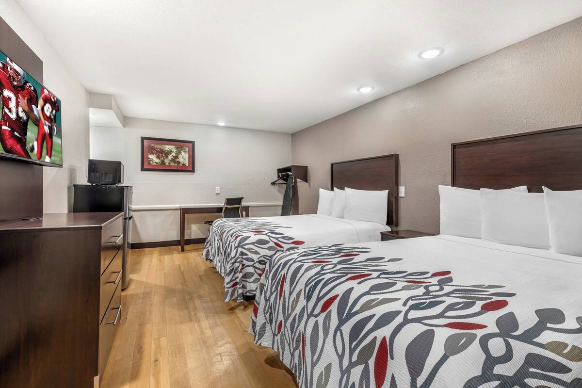 A hotel room with a long bed, a desk, a chair, a television, and a framed picture on the wall.