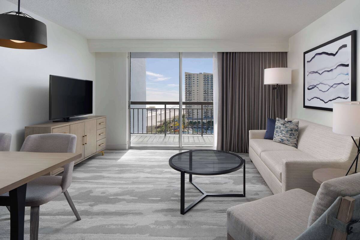A modern hotel room with a balcony, a flat-screen TV, a dining table, and a sofa.