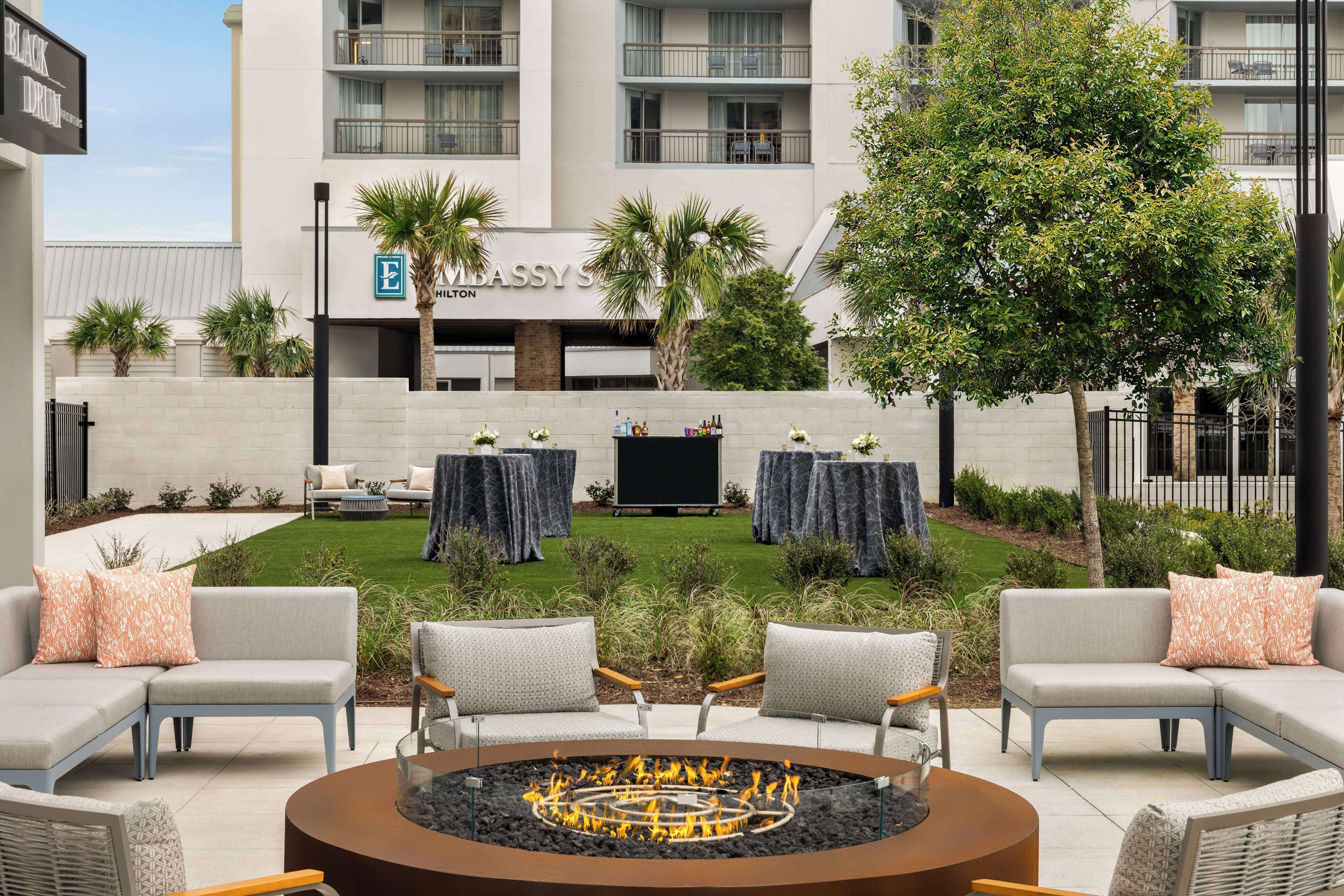 An outdoor seating area with a fire pit, sofas, and tables, surrounded by a well-maintained lawn and palm trees.