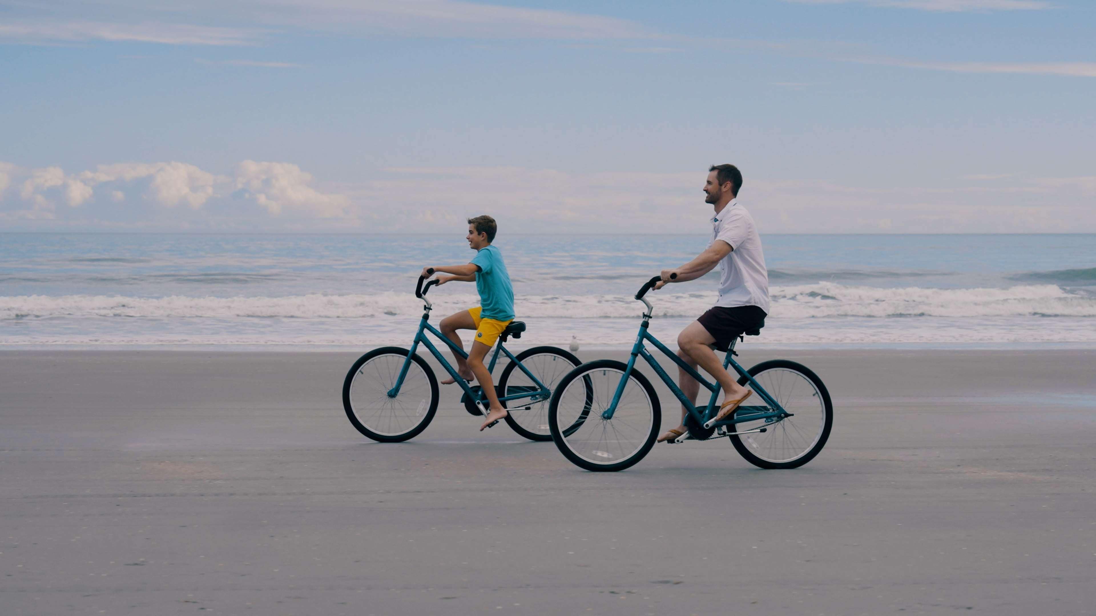 Dos personas montando en bicicleta sobre la playa, con el océano al fondo.