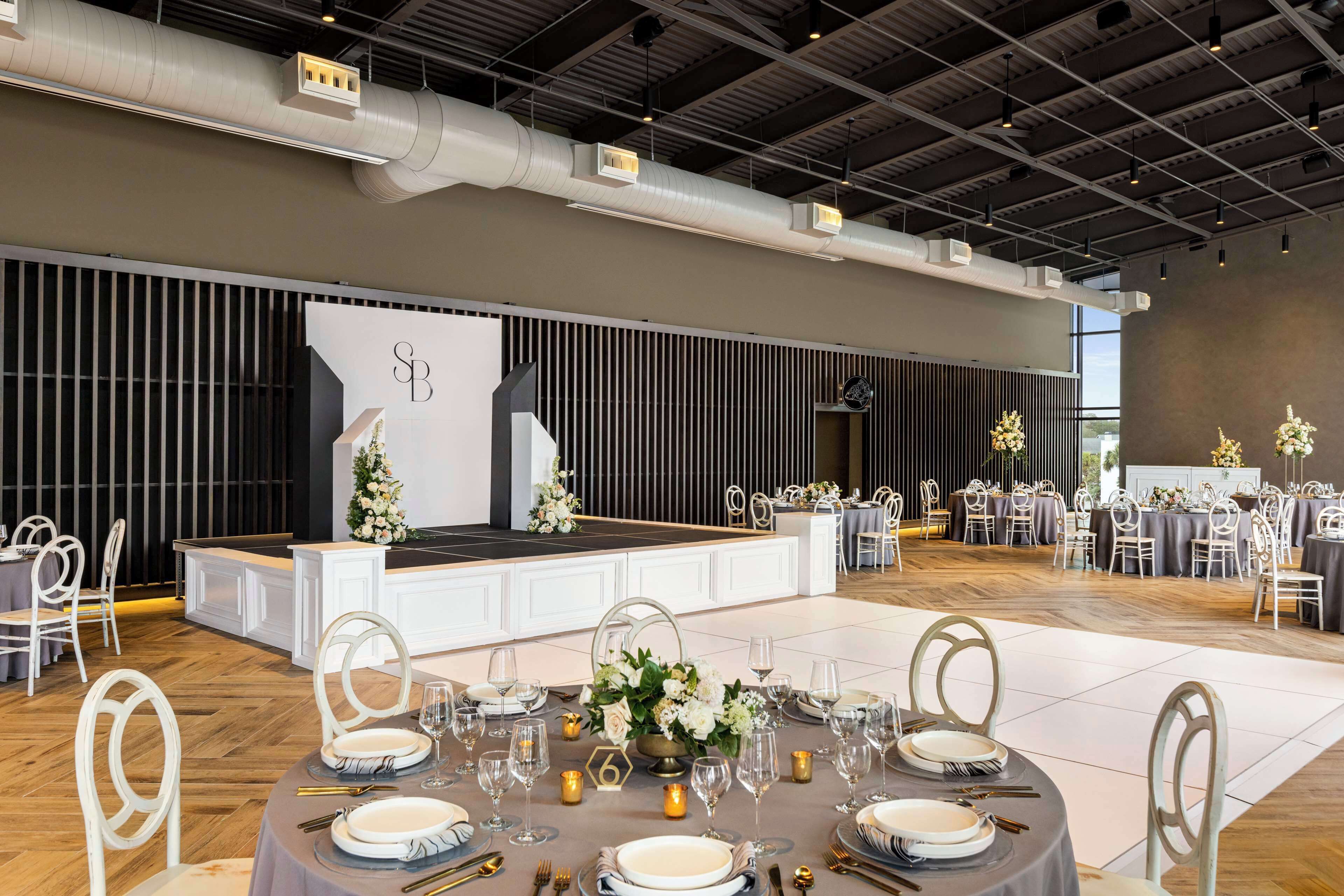 Un salón de banquetes con mesas redondas dispuestas para un evento formal, con sillas blancas y un escenario con un telón de fondo.