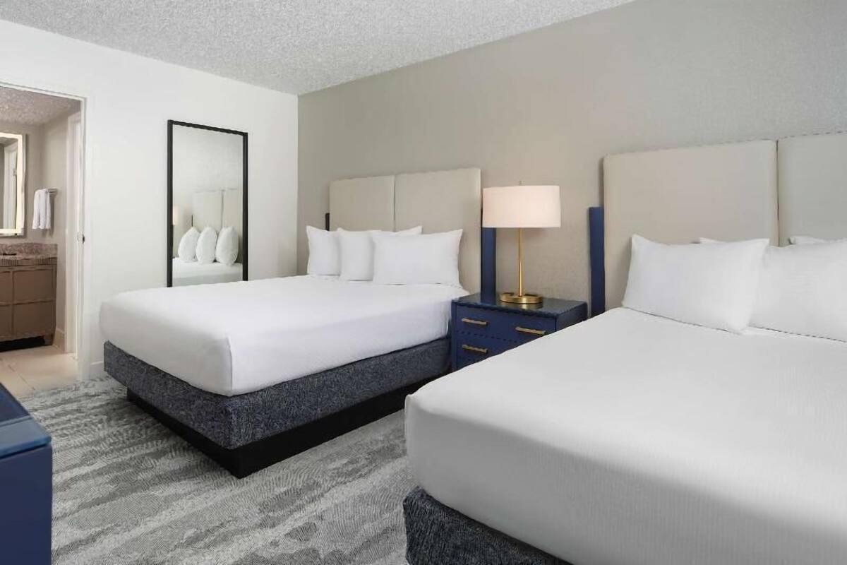 A hotel room with two beds, a nightstand with a lamp, a mirror, and a bathroom visible through an open door.