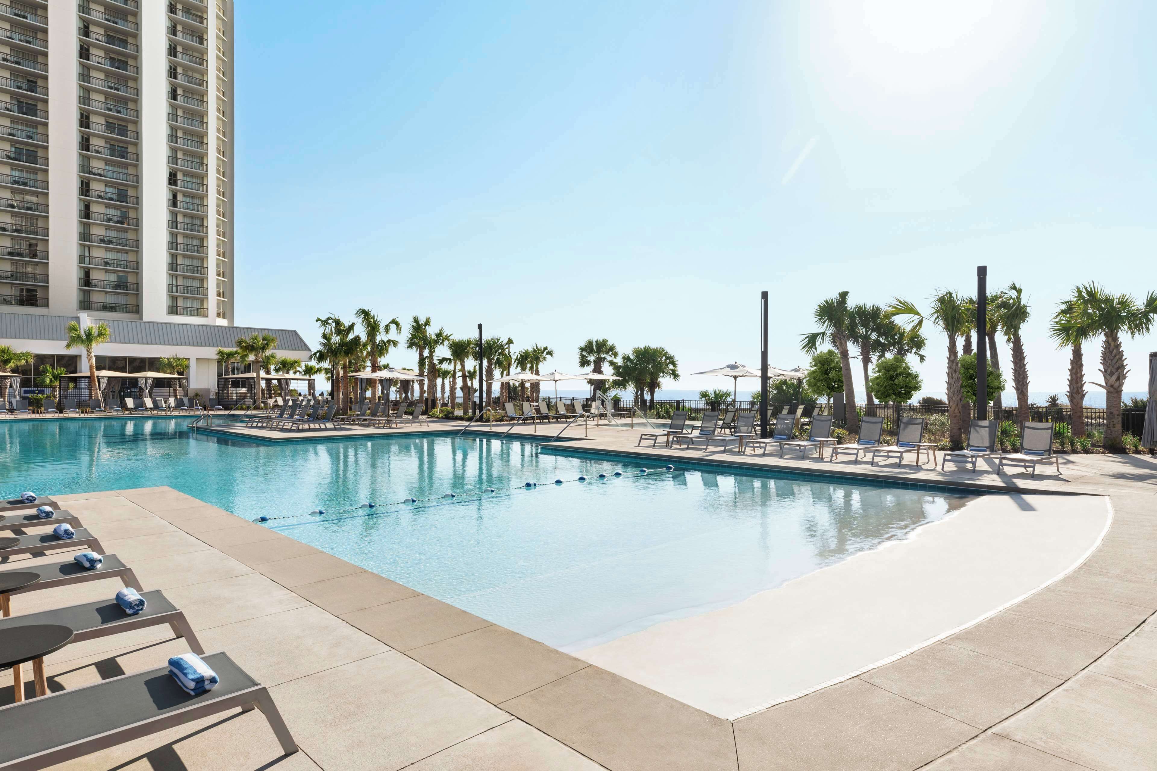 A hotel pool area with lounge chairs and umbrellas, surrounded by palm trees and a tall building in the background.