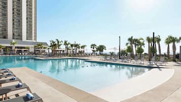 A hotel pool area with lounge chairs and umbrellas, surrounded by palm trees and a tall building in the background.